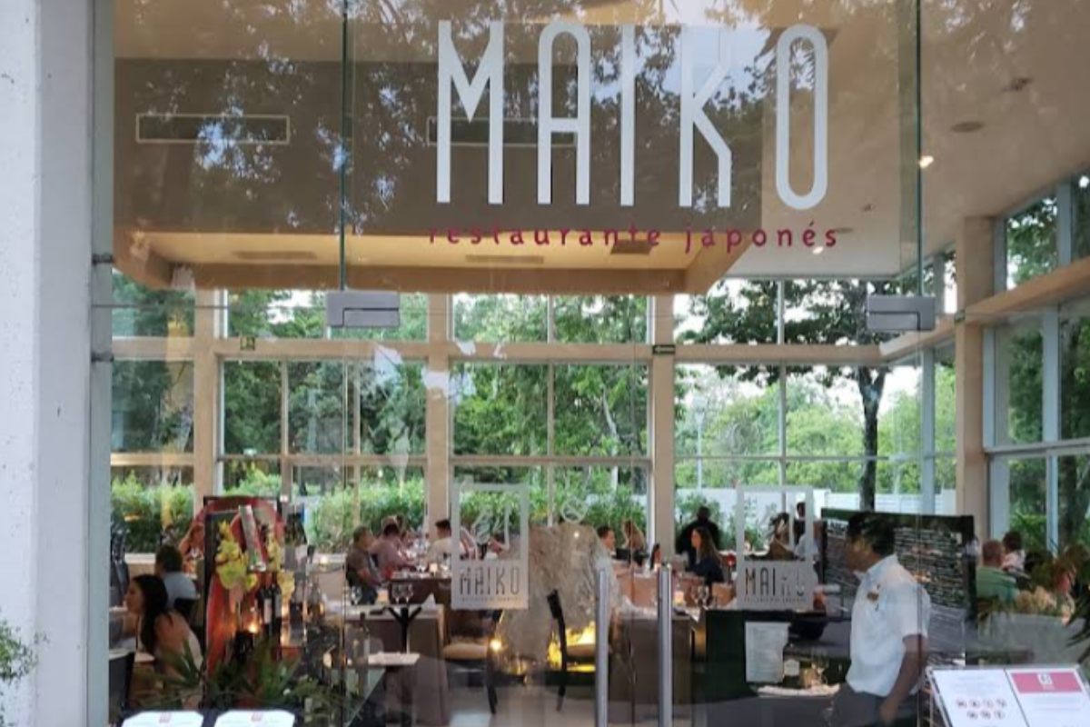 Maiko Japanese Restaurant entrance with large glass windows, tropical greenery reflection, and the MAIKO sign above the dining room inside Tulum Country Club