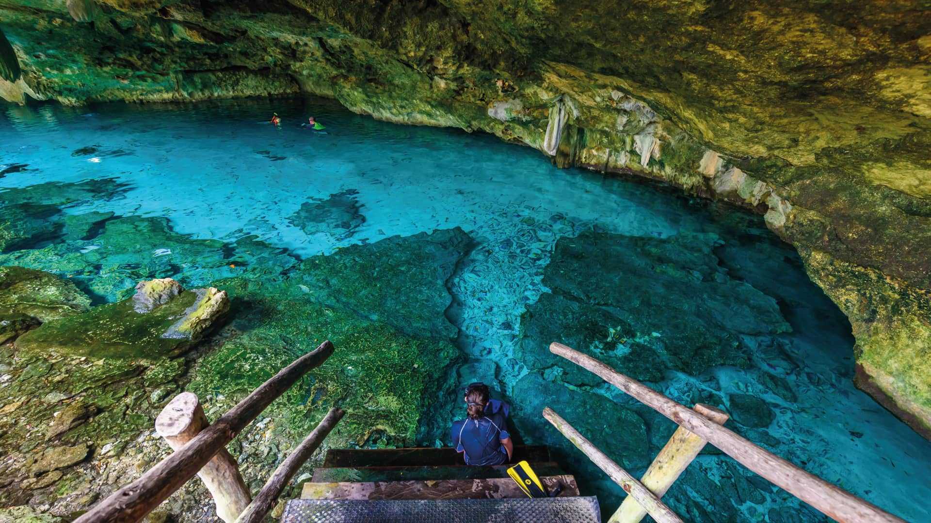 Snorkeler descending wooden stairs into the turquoise cavern pool of Dos Ojos cenote near Akumal