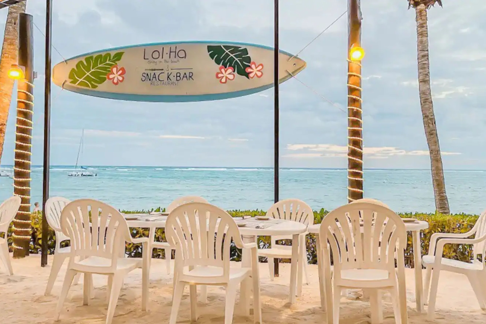 Lol-Ha beachfront snack bar with surfboard sign, white plastic chairs, and turquoise Caribbean Sea in the background
