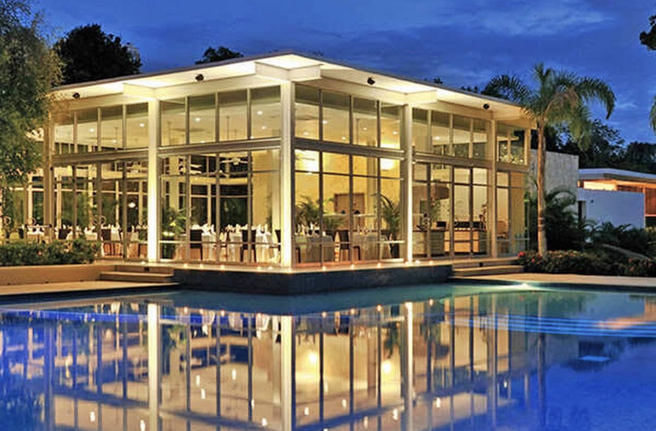 Modern glass-walled Alux Gourmet restaurant at dusk, reflecting on a tranquil pool inside Tulum Country Club