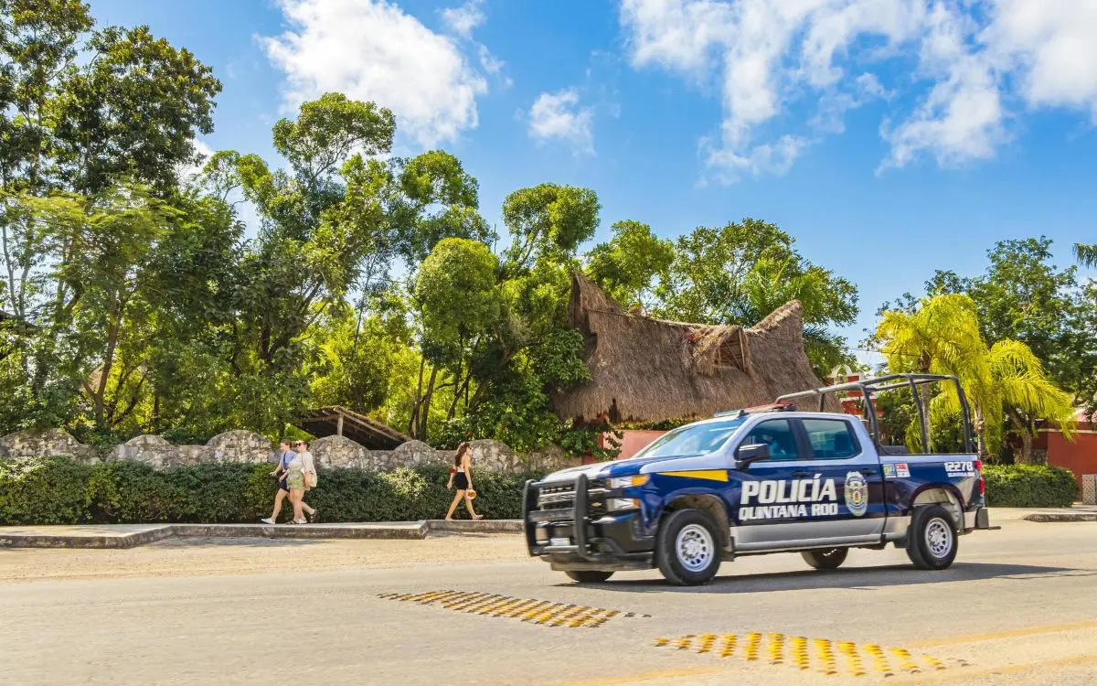 A Policía Quintana Roo patrol truck driving past a thatched-roof palapa and lush greenery in Akumal, with visitors walking safely along the sidewalk