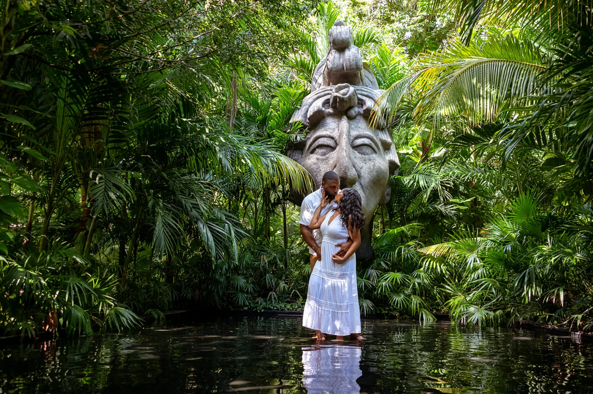 Couple kissing in a jungle cenote beneath a Mayan stone sculpture during an engagement session