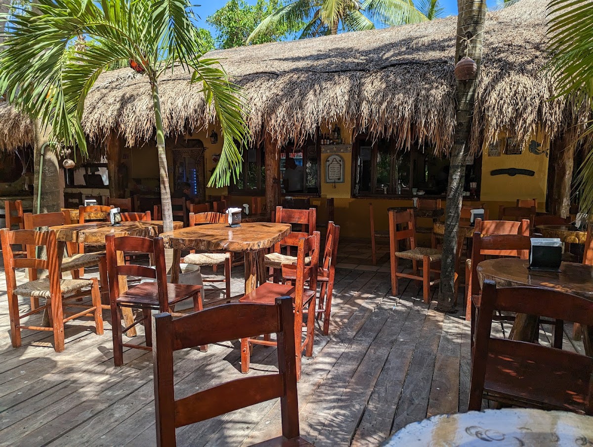 La Cueva del Pescador open-air beachfront dining area with palapa roof, wooden tables, chairs, and palm trees